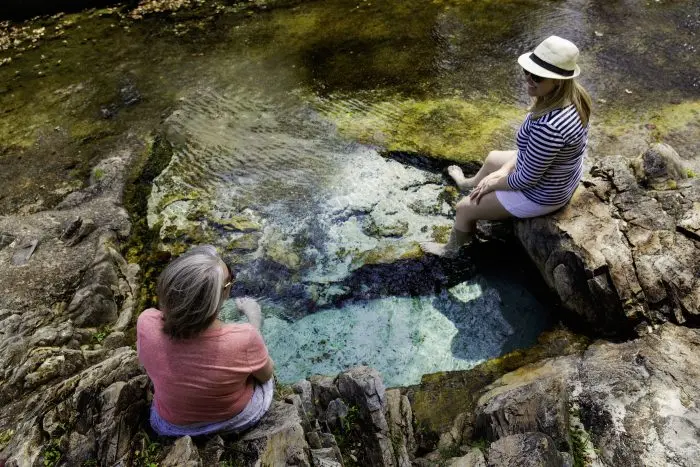 Two people sit on rocks soaking feet in a clear spring pool outdoors