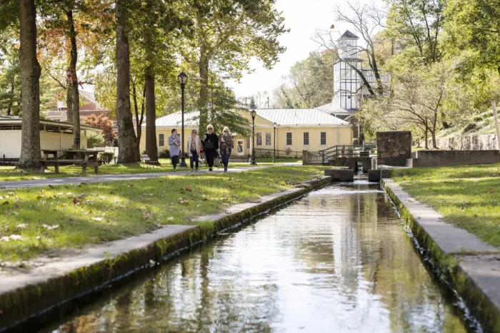 People walk along a canal in a park with historic buildings and trees
