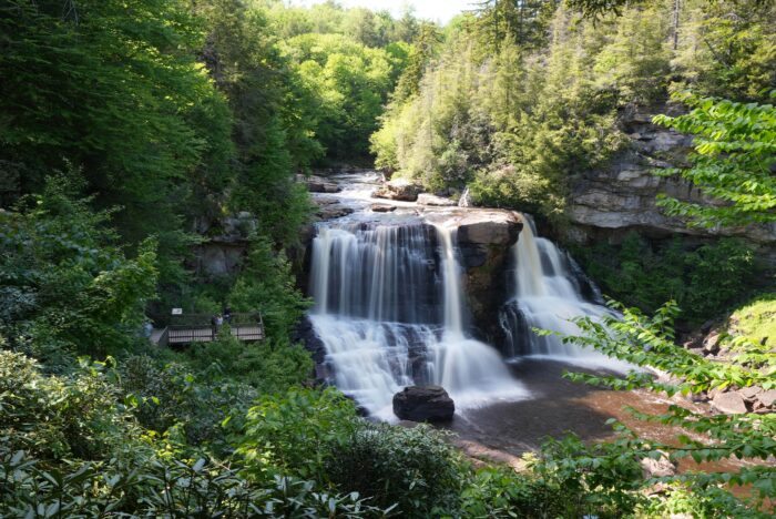 Powerful Blackwater Falls plunges into a rocky basin surrounded by thick forest, with a wooden viewing platform visible on the left.
