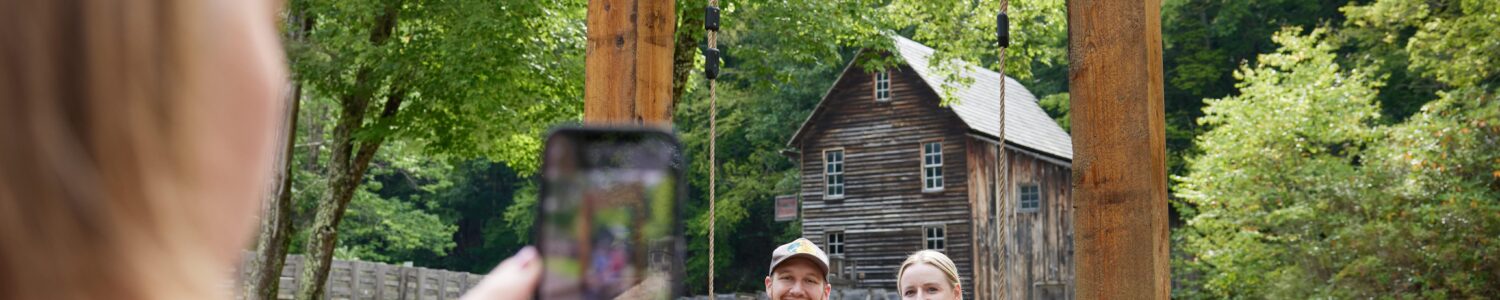 Couple on a rustic “Almost Heaven” swing at Babcock State Park with Glade Creek Grist Mill in background poses for a picture