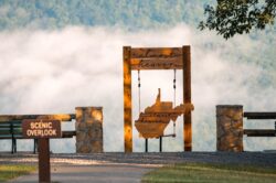 "Almost Heaven" swing featuring a wooden cutout of West Virginia at Pipestem Resort State Park, overlooking a scenic mountain valley with early morning fog — a top West Virginia overlook for photography, sightseeing, and outdoor travel.