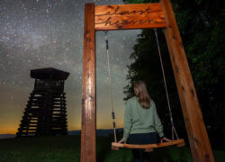 Woman standing beside an "Almost Heaven" swing at Droop Mountain Battlefield State Park in West Virginia, with a historic wooden lookout tower and a star-filled night sky — a scenic spot for stargazing, photography, and outdoor adventure.
