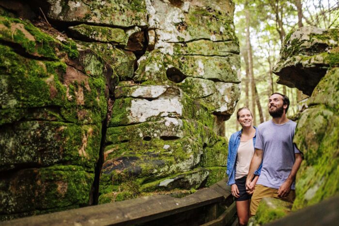 A smiling couple walks along a wooden trail surrounded by large moss-covered rock formations.