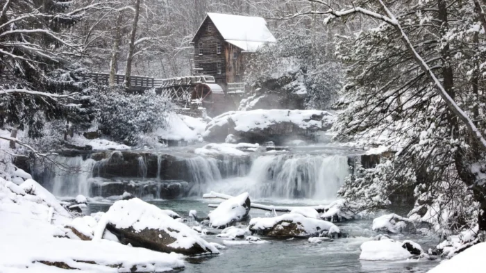 Snow-covered waterfall and stream flowing past a historic wooden mill in a winter forest.
