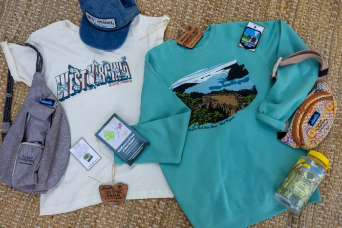 West Virginia State Parks apparel and accessories arranged on woven mat, including T-shirts, sweatshirt, hat, sling bags and water bottle.