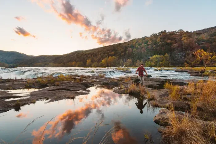 Person standing on rocky riverbank at sunset with reflections in still water.