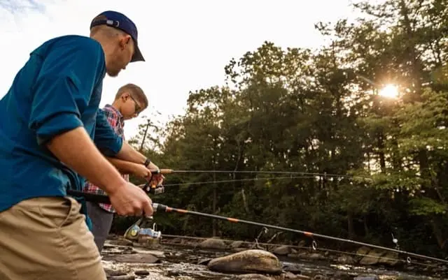 Two people stand on rocks along a shallow riverbank, casting fishing lines into the water as the sun sets behind a line of trees.