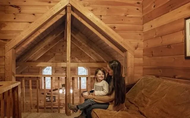 A mother and child sit together on a couch in a warm, wooden cabin loft, sharing a quiet moment beneath a pitched ceiling made of pine beams.