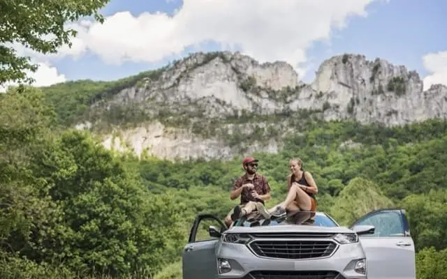 A couple sits on the hood of a silver SUV parked in front of Seneca Rocks in West Virginia, enjoying snacks with the towering rock formation in the background.