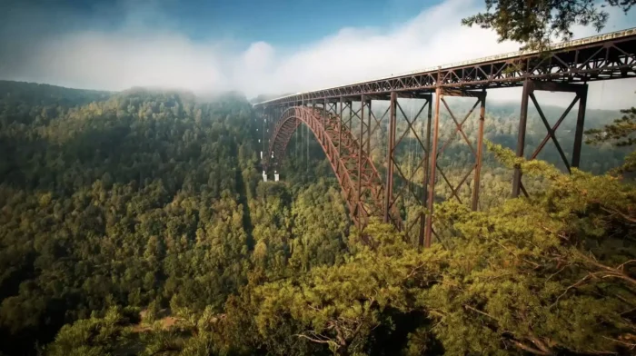 Steel arch bridge spanning a forested gorge in morning mist.