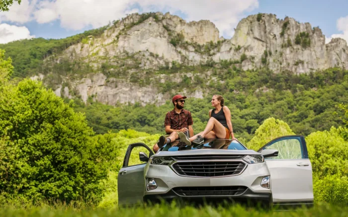 Two people sitting on car with open doors in front of forested mountains