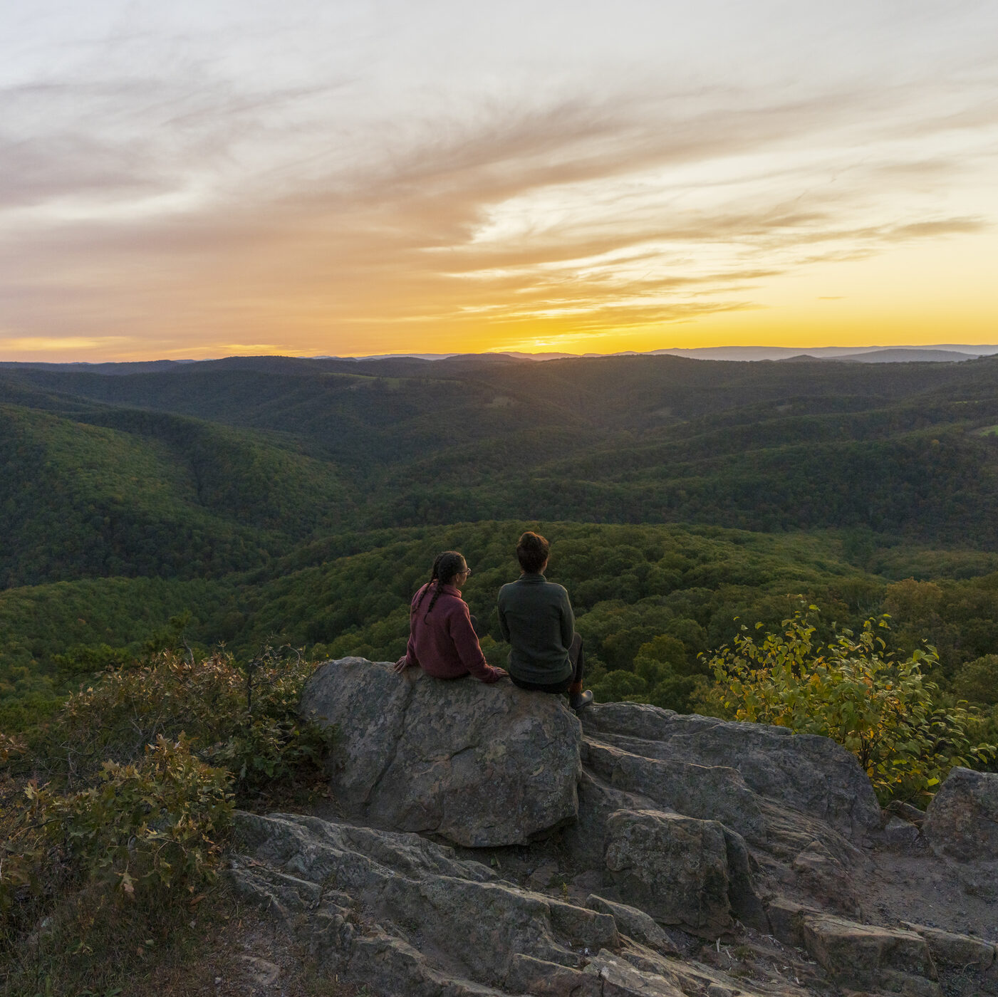 Two people sitting on a rock overlooking a forested valley at sunset with a colorful sky.